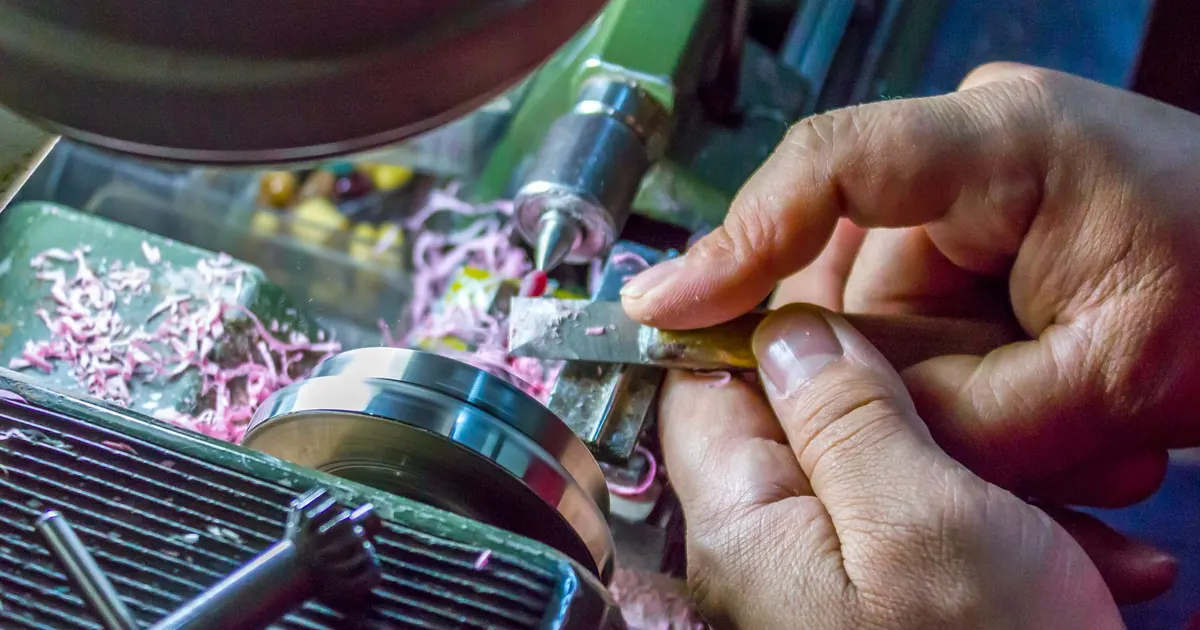 Hands holding a small hand tool against a part in a lathe with swarf in the tray beside it